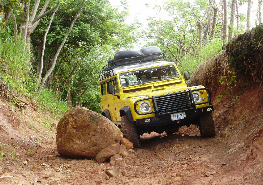 Ein gelber Geländewagen auf einer Schotterpiste unterwegs, wobei einem großen Stein ausgewichen werden muss, der auf den Weg gefallen ist.