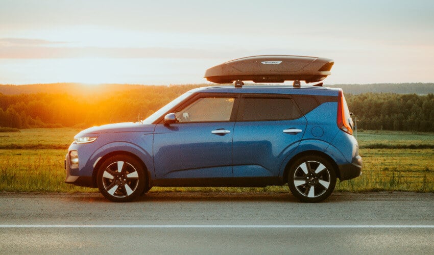 Ein blaues Auto mit einer Dachbox steht am Straßenrand und im Hintergrund ein Sonnenaufgang.