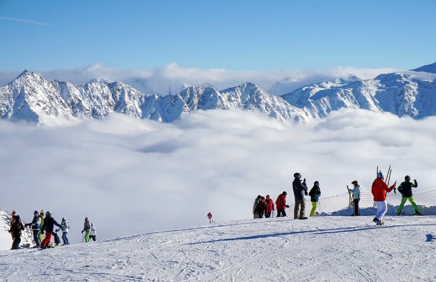 Blick von der Skipiste Richtung wolkenbedecktes Tal. Am Berg strahlend blauer Himmel.